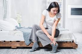 download (24) mulher sentada na cama segurando o tornozelo com expressão de dor matinal.